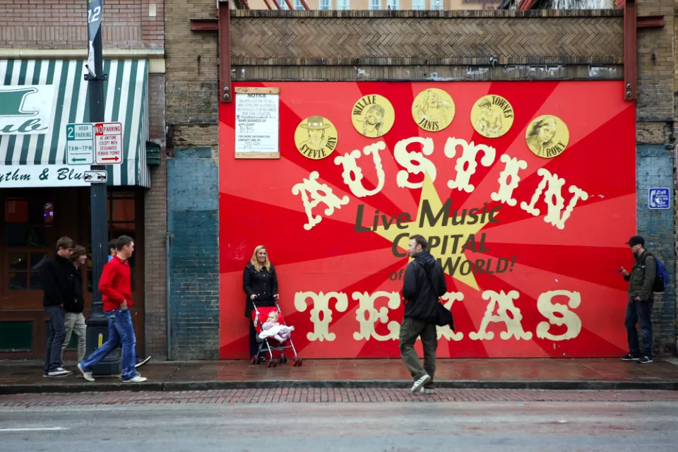 A woman with a baby in a stroller and two men are milling around in front a sign that reads Austin Texas: Live Music Capital of the World.